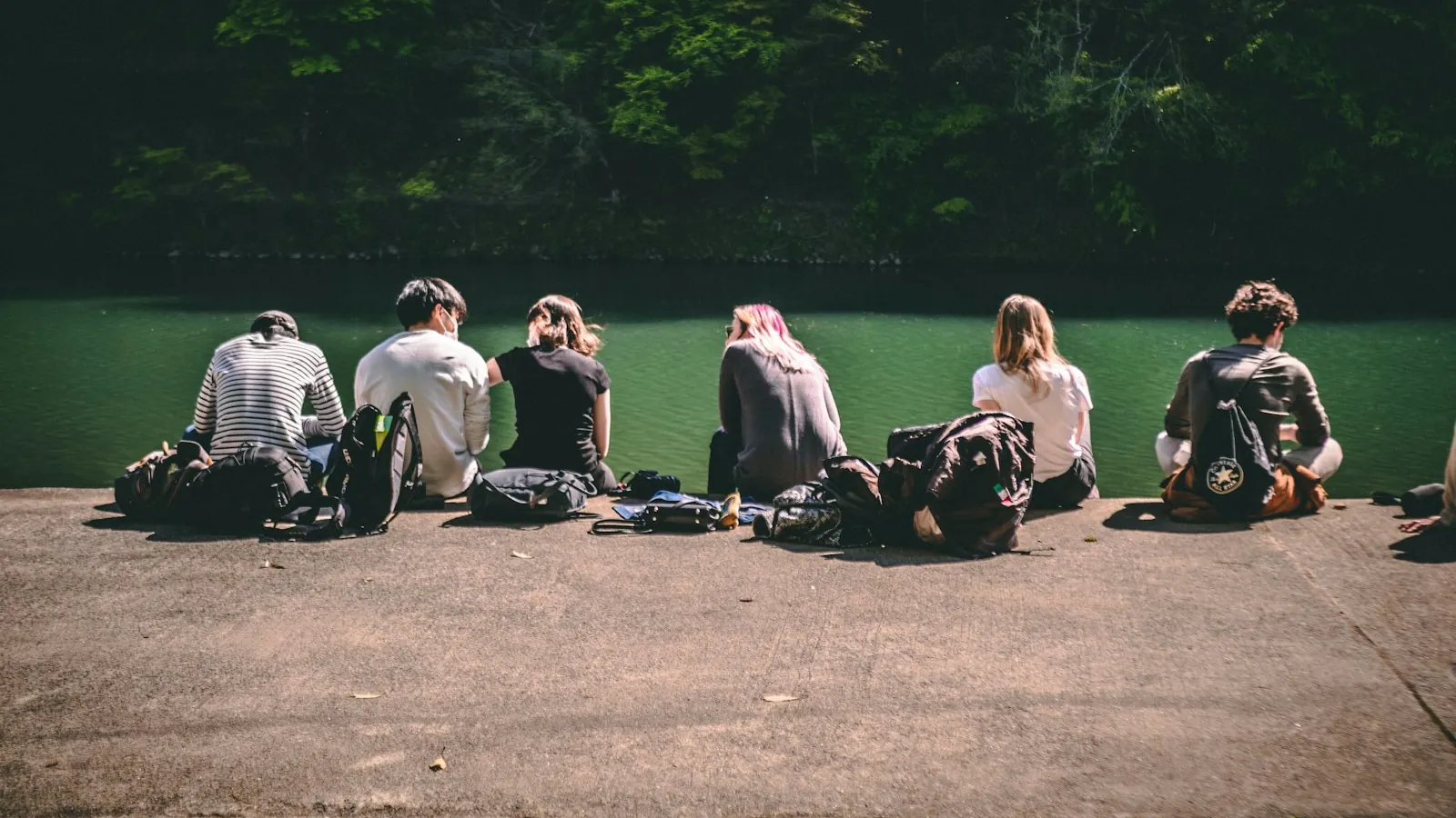 Gruppe unge mennesker sitter sammen og snakker ved en elvebredd i sol